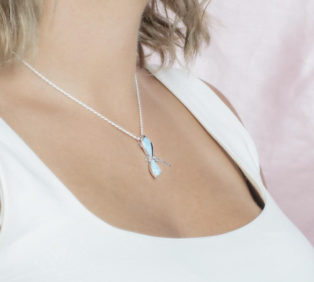 In this photo there is a model turned to the right with blonde hair and a white shirt, wearing a sterling silver dragonfly pendant with blue larimar gemstones.