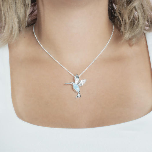 In this photo there is a model with blonde hair and a white shirt, wearing a sterling silver flying hummingbird pendant with topaz and one blue larimar gemstone.