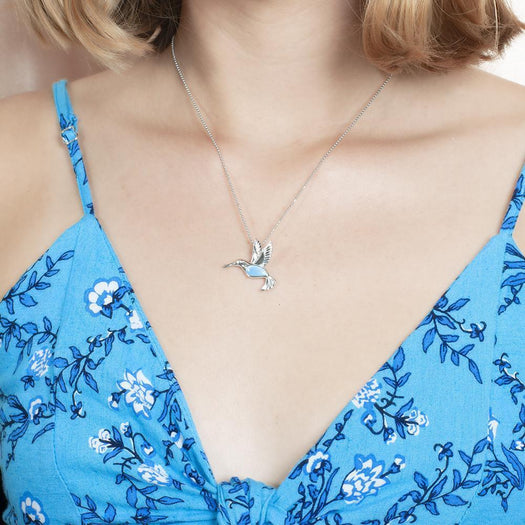 In this photo there is a model with blonde hair and a blue dress with flowers, wearing a sterling silver hummingbird pendant with one blue larimar gemstone.