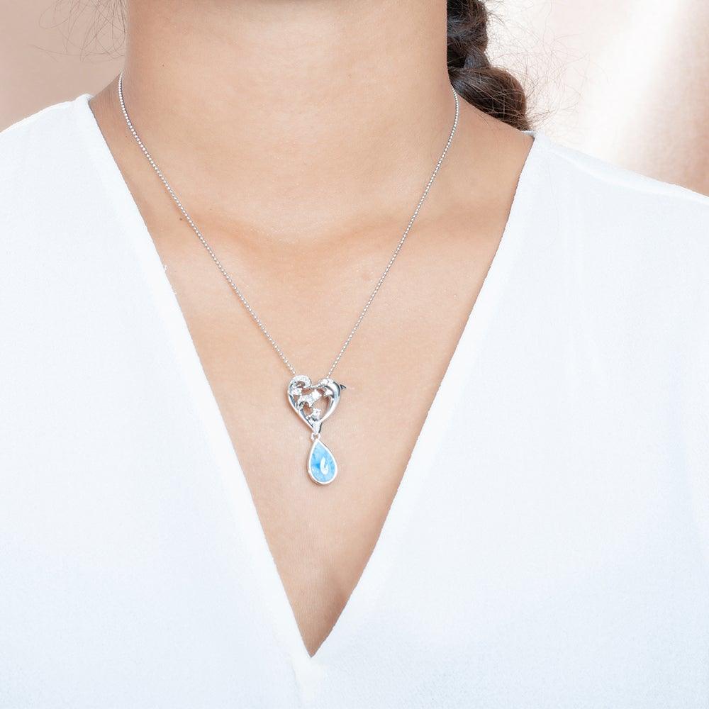 The picture shows a model wearing a sterling silver larimar dolphin pendant with a heart and wave motif, aquamarines, and topaz.