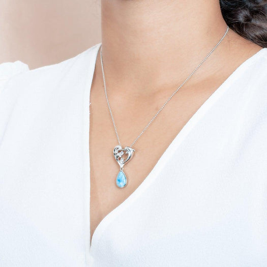 The picture shows a model wearing a sterling silver larimar dolphin pendant with a heart and wave motif, aquamarines, and topaz.