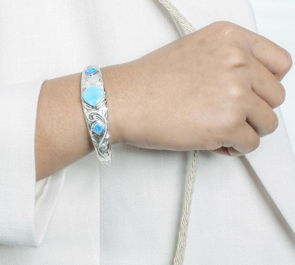 The picture shows a model wearing a 925 sterling silver bangle with one larimar gemstone centered by two opalite gemstones with hand-engravings.