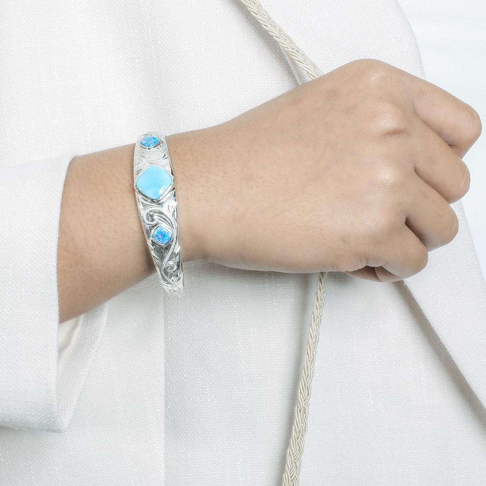 The picture shows a model wearing a 925 sterling silver bangle with one larimar gemstone centered by two opalite gemstones with hand-engravings.