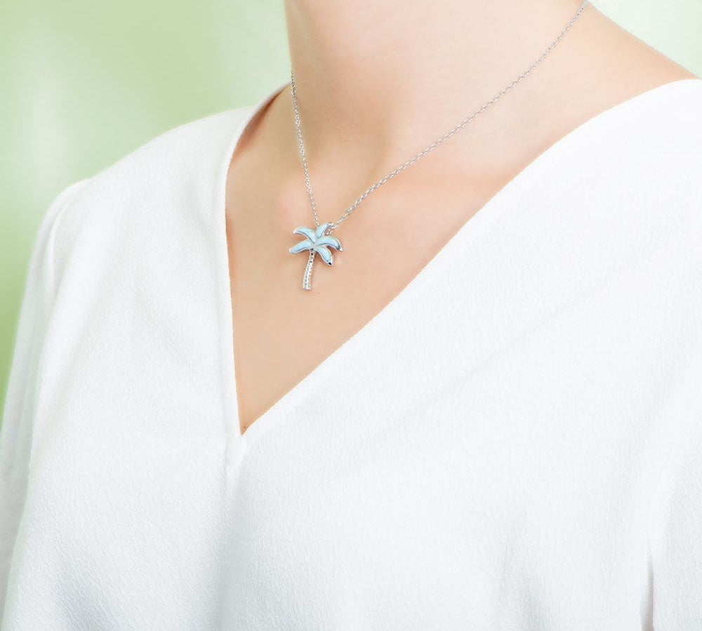 In this picture there is a model faced to the left with a white shirt, wearing a palm tree pendant with blue larimar gemstones set in sterling silver.