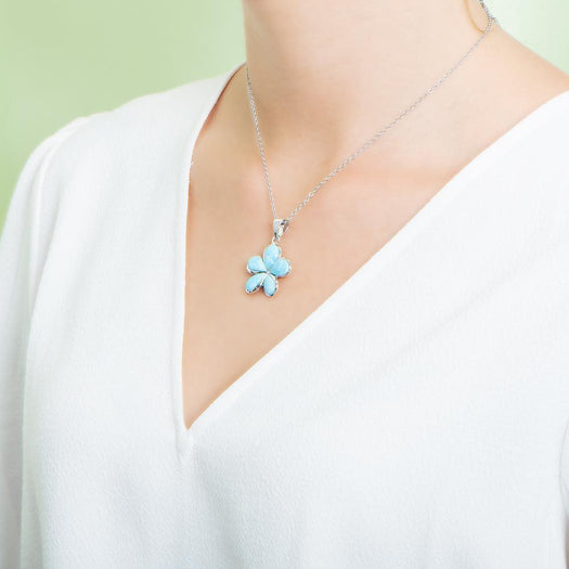 In this photo there is a model with a white shirt turned to the left, wearing a medium sized sterling silver plumeria pendant with blue larimar gemstones.