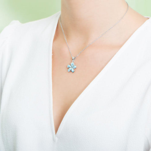 In this photo there is a model with a white shirt turned to the left, wearing a small sterling silver plumeria pendant with blue larimar gemstones.
