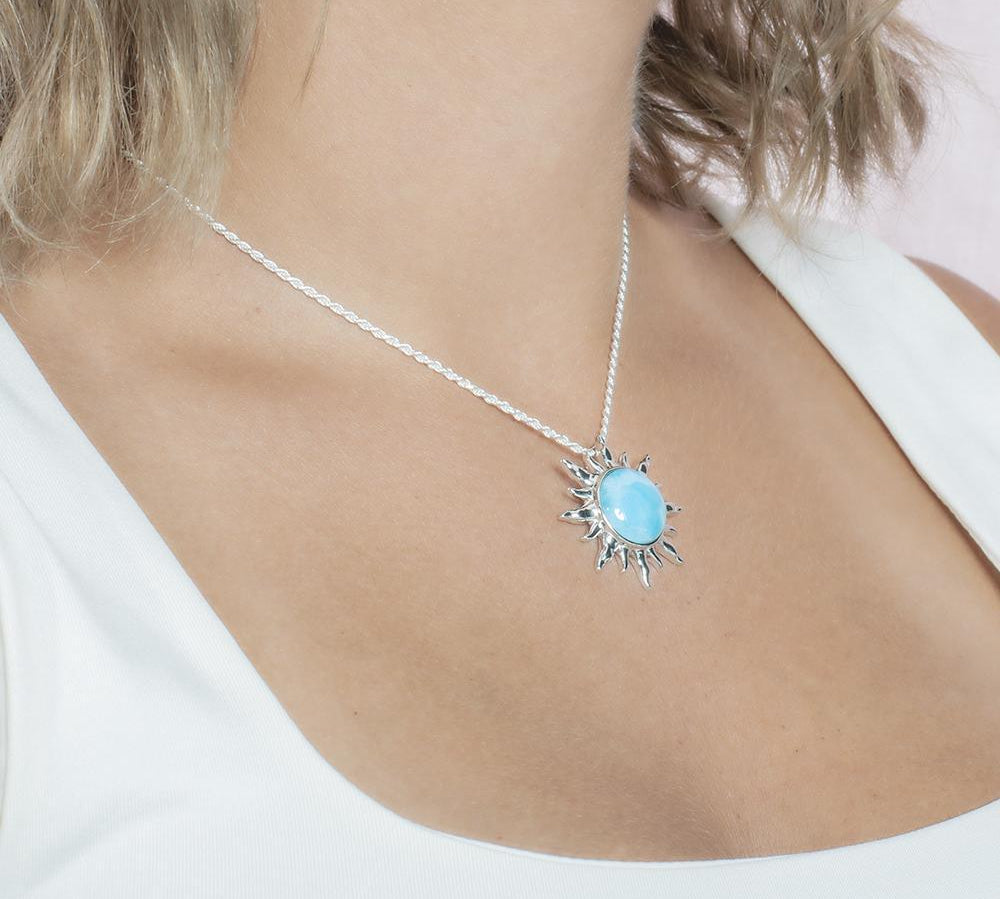 In this picture there is a model with blonde hair and a white shirt turned to the right wearing a sun pendant with a blue larimar gemstone set in sterling silver.