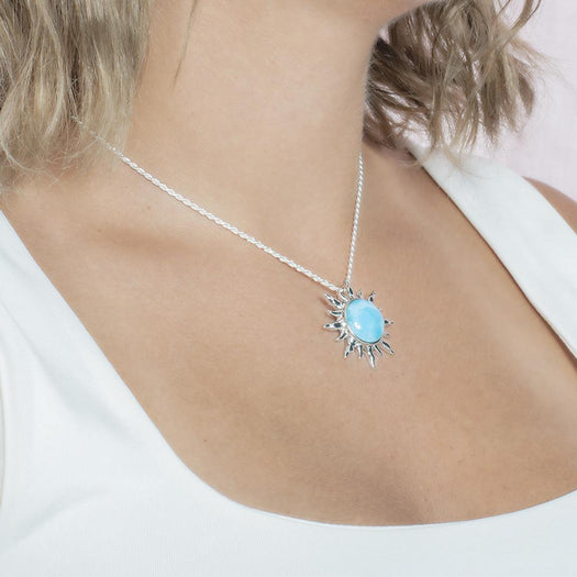 In this picture there is a model with blonde hair and a white shirt turned to the right wearing a sun pendant with a blue larimar gemstone set in sterling silver.