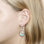 The picture shows a model wearing a 925 sterling silver larimar whale wave lever-back earring with topaz.