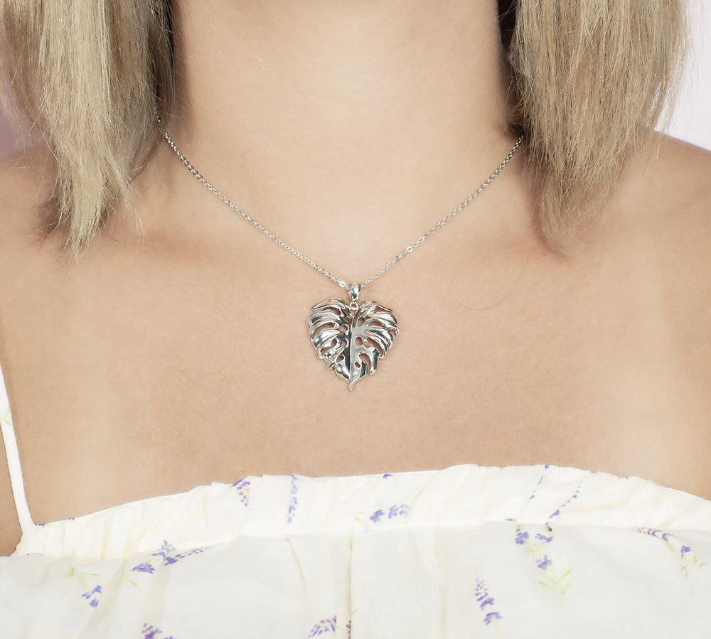 In this photo there is a model with blonde hair and white shirt with flowers, wearing a sterling silver monstera pendant.