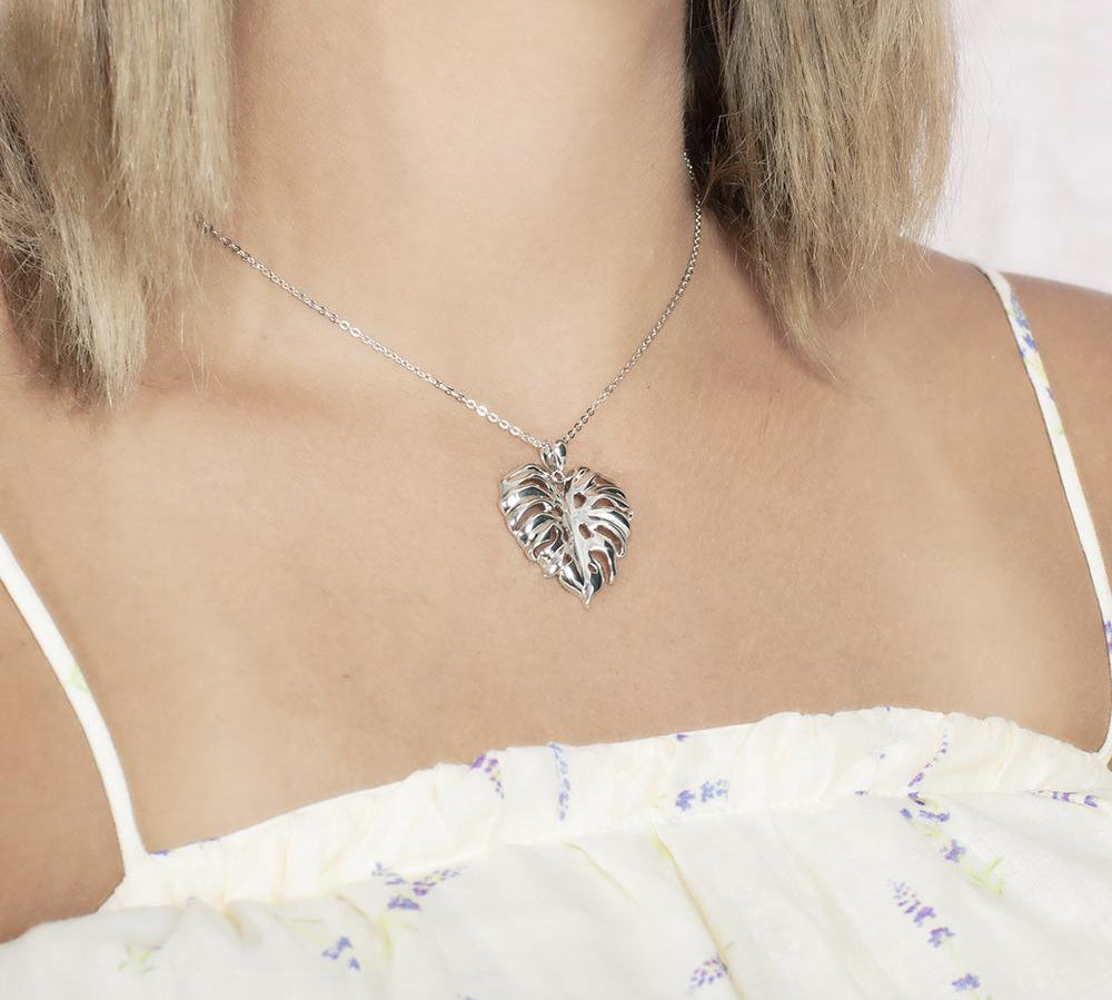 In this photo there is a model with blonde hair and white shirt with flowers turned to the right, wearing a sterling silver monstera pendant.