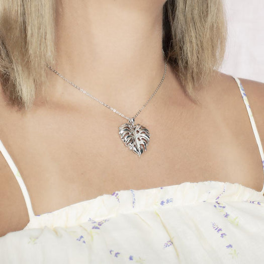 In this photo there is a model with blonde hair and white shirt with flowers turned to the right, wearing a sterling silver monstera pendant.