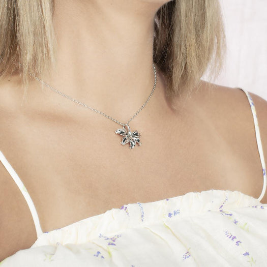 In this photo there is a model turned to the right with blonde hair and a white shirt with purple flowers, wearing a sterling silver naupaka flower pendant with cubic zirconia.
