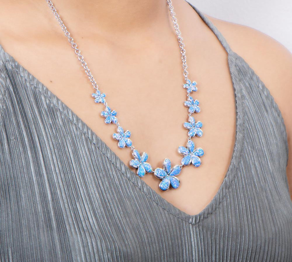 In this photo there is a model with a gray shirt wearing a sterling silver necklace with nine plumeria flowers and blue opalite gemstones.
