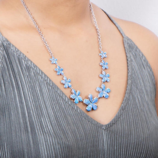 In this photo there is a model with a gray shirt wearing a sterling silver necklace with nine plumeria flowers and blue opalite gemstones.