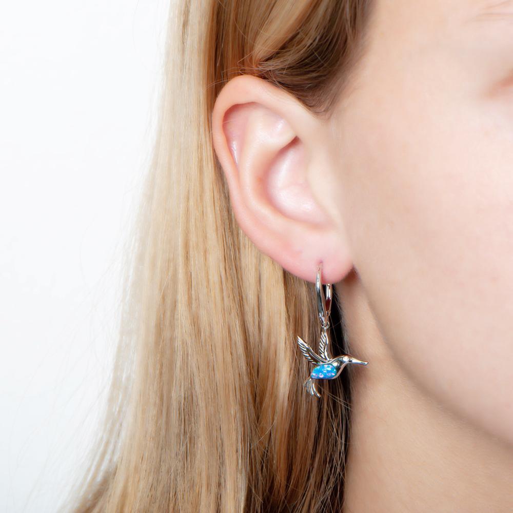 In this photo there is a model with blonde hair wearing sterling silver hummingbird dangle earrings with blue opalite and topaz gemstones.