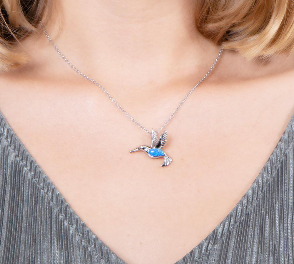 In this photo there is a model with blonde hair and a gray shirt, wearing a sterling silver hummingbird pendant with topaz and blue opalite gemstones.