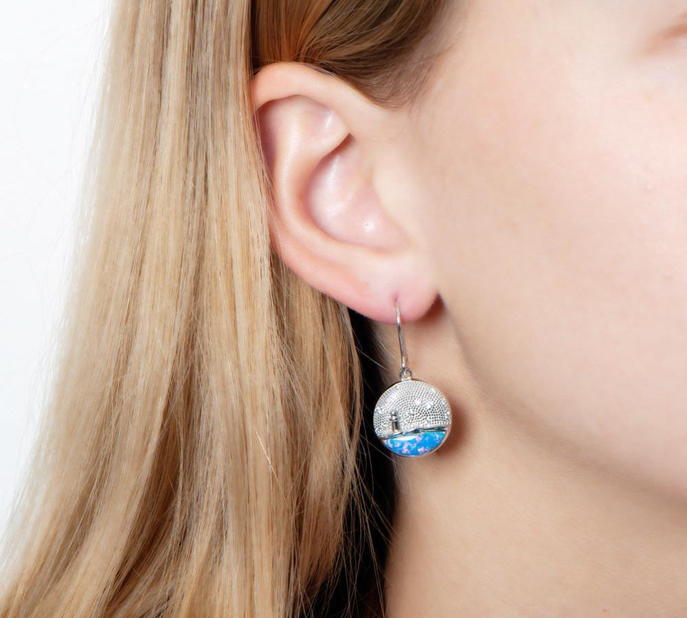 In this photo there is a close-up of a model with blonde hair wearing sterling silver lighthouse medallion hook earrings with blue opalite and topaz gemstones.