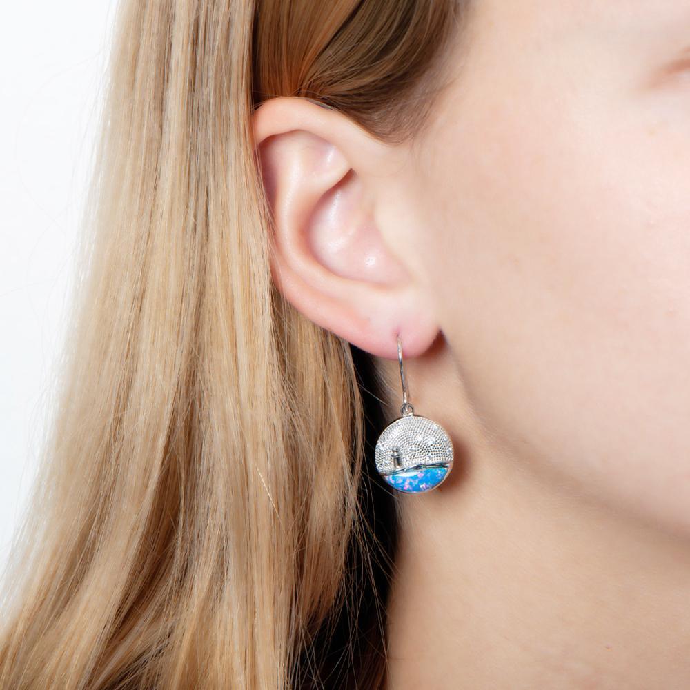 In this photo there is a close-up of a model with blonde hair wearing sterling silver lighthouse medallion hook earrings with blue opalite and topaz gemstones.