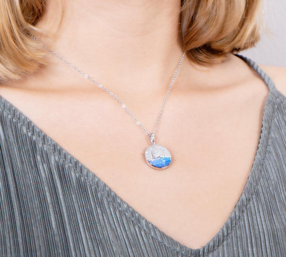 In this photo there is a model turned slightly to the right with blonde hair and a gray shirt, wearing a sterling silver lighthouse medallion pendant with blue opalite and topaz gemstones.