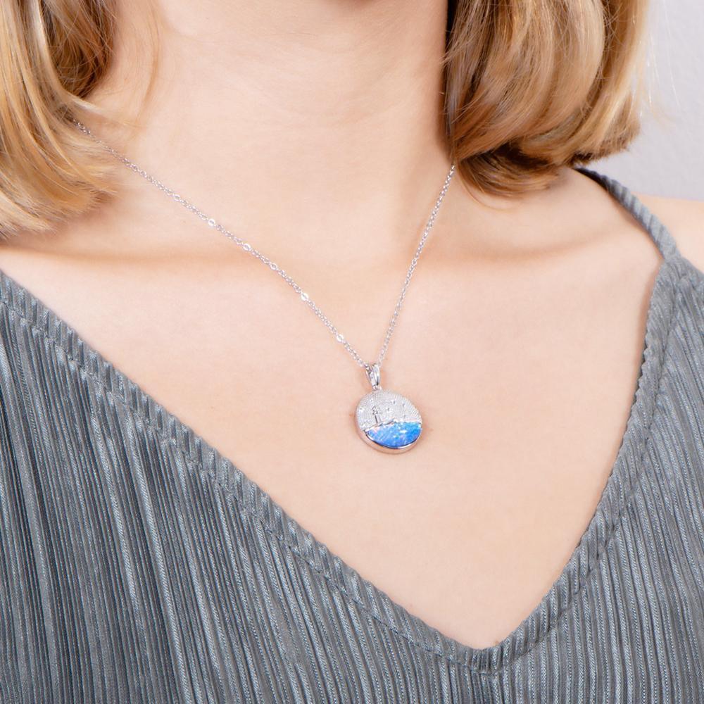 In this photo there is a model turned slightly to the right with blonde hair and a gray shirt, wearing a sterling silver lighthouse medallion pendant with blue opalite and topaz gemstones.