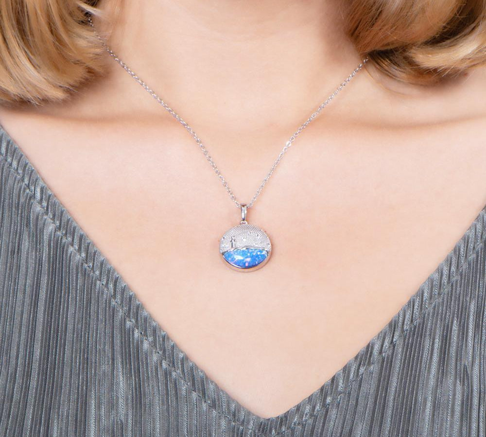 In this photo there is a model with blonde hair and a gray shirt, wearing a sterling silver lighthouse medallion pendant with blue opalite and topaz gemstones.