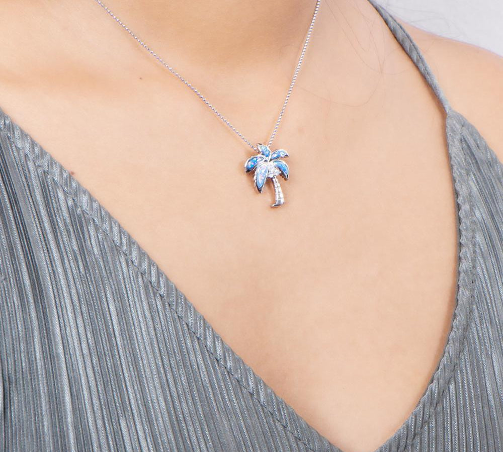 In this photo there is a model with a gray shirt turned slightly to the right, wearing a sterling silver palm tree pendant with blue opalite and three topaz gemstones.