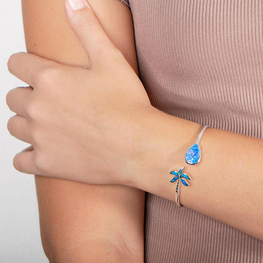 In this photo there is a close up of a model with a beige shirt, wearing a sterling silver palm tree and teardrop bangle with blue opalite gemstones.