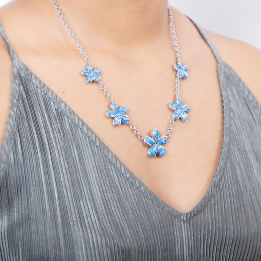 In this photo there is a model with a gray shirt wearing a sterling silver charm necklace with five plumeria flowers and blue opalite gemstones.