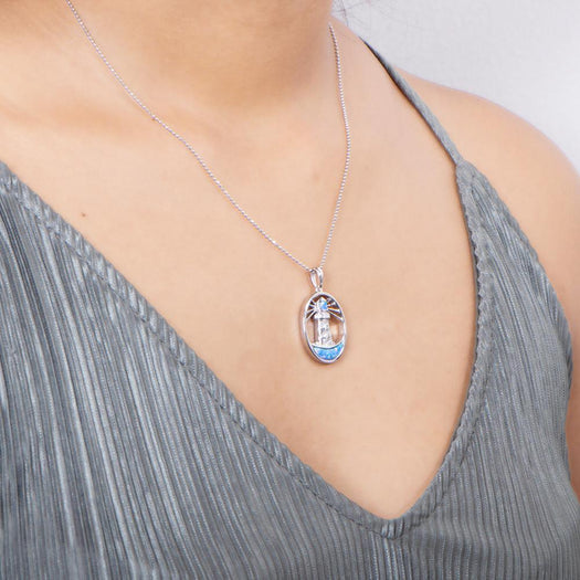 In this photo there is a model with a gray shirt turned slightly to the right, wearing a sterling silver shining lighthouse pendant with blue opalite and topaz gemstones.