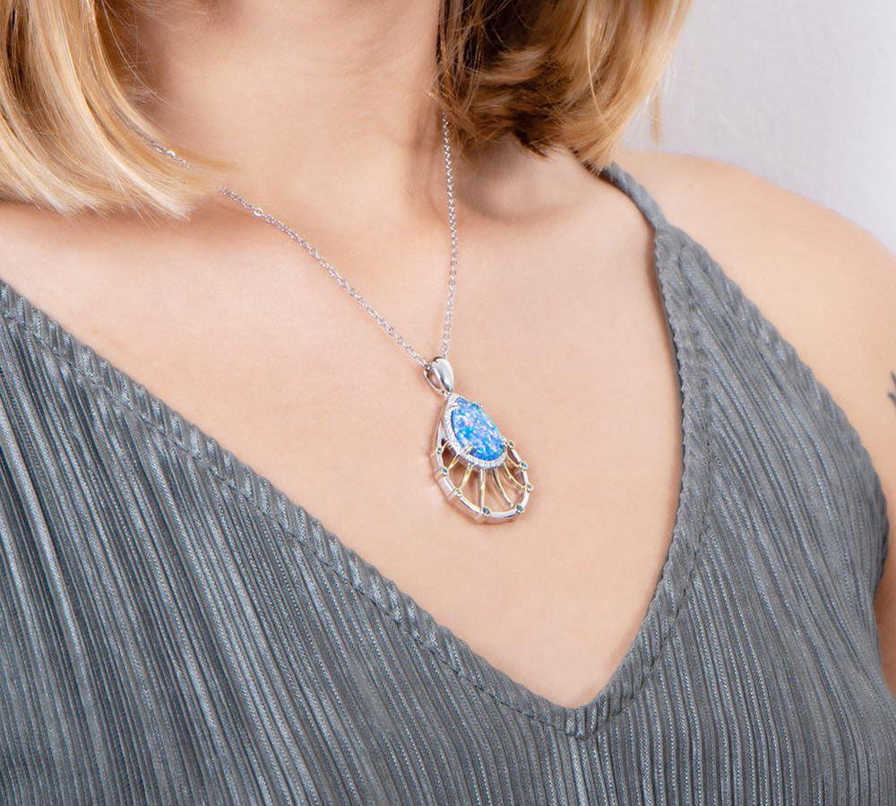 In this photo there is a model turned slightly to the right with blonde hair and a gray shirt, wearing a sterling silver and gold plated teardrop pendant with blue opalite, aquamarine, and topaz gemstones.