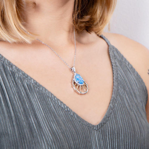 In this photo there is a model turned slightly to the right with blonde hair and a gray shirt, wearing a sterling silver and gold plated teardrop pendant with blue opalite, aquamarine, and topaz gemstones.