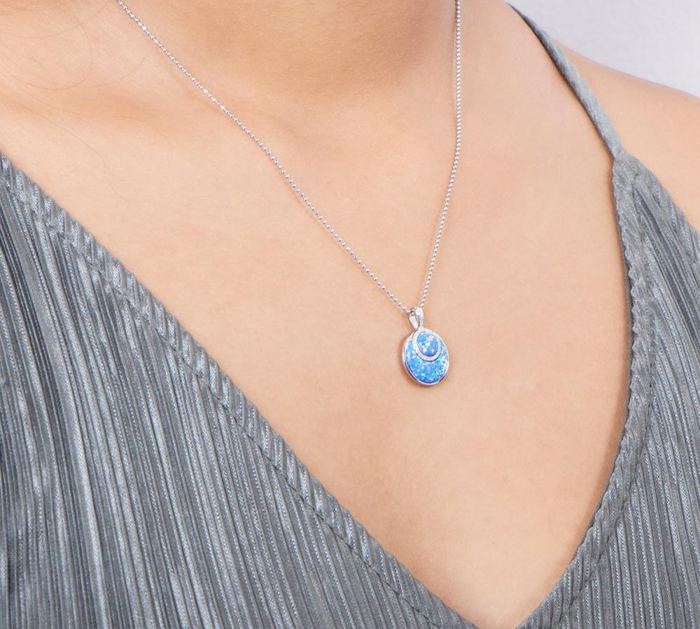 In this photo there is a model with a gray shirt turned slightly to the right, wearing a sterling silver circle pendant with blue opalite and topaz gemstones.