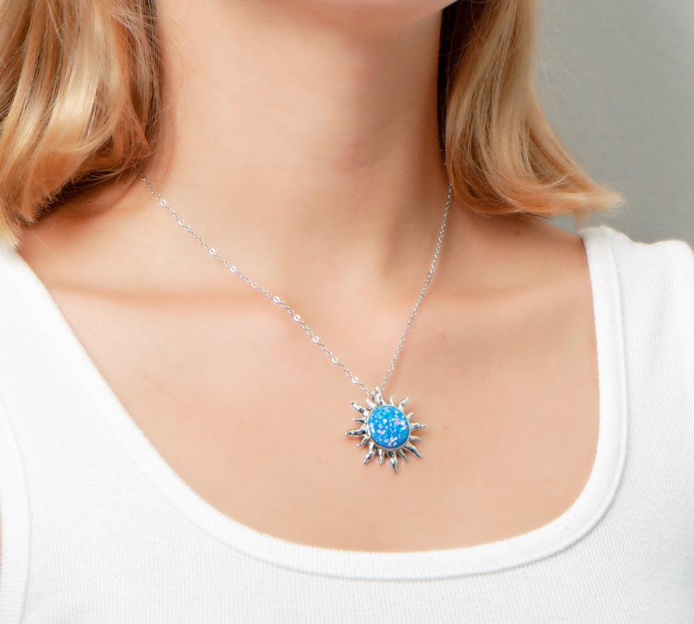 In this photo there is a model turned slightly to the right with blonde hair and a white shirt, wearing a sterling silver sun pendant with one blue opalite gemstone.