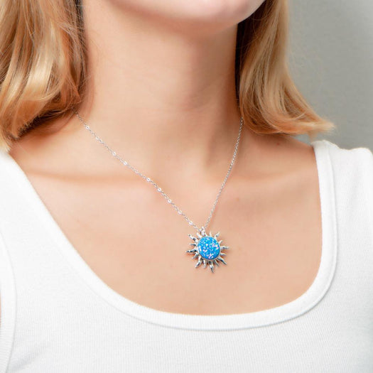 In this photo there is a model turned slightly to the right with blonde hair and a white shirt, wearing a sterling silver sun pendant with one blue opalite gemstone.