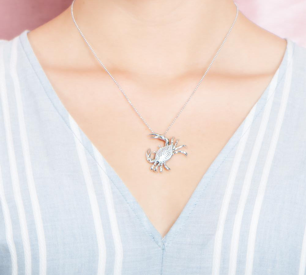 The picture shows a model wearing a 925 sterling silver, white gold plated, pavé blue crab pendant with cubic zirconia.