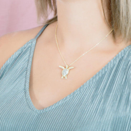 In this photo there is a model with blonde hair and a teal shirt turned to the left, wearing a yellow gold sea turtle pendant with cubic zirconia.