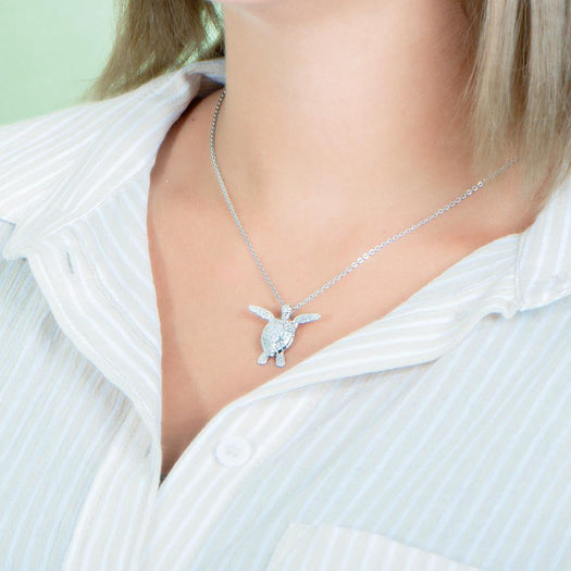 In this photo there is a model turned to the left with blonde hair and a tan and white striped shirt, wearing a white gold sea turtle pendant with cubic zirconia.