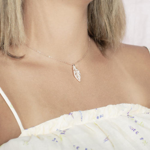 In this photo there is a model turned to the right with blonde hair and a white shirt with purple flowers, wearing a rose gold maile leaf pendant with topaz gemstones.