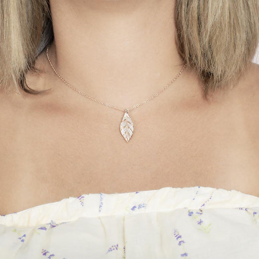 In this photo there is a model with blonde hair and a white shirt with purple flowers, wearing a rose gold maile leaf pendant with topaz gemstones.