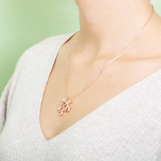 In this photo there is a model turned to the left with blonde hair and a white shirt, wearing a rose gold sea turtle pendant with topaz gemstones.