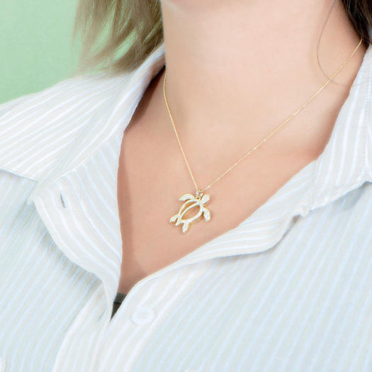 In this photo there is a model turned to the left with blonde hair and a tan and white striped shirt, wearing a yellow gold sea turtle pendant with topaz gemstones.