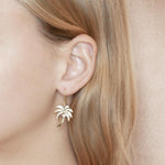 In this photo is a close-up of a model with blonde hair wearing a pair of yellow gold palm tree hook earrings with topaz gemstones.