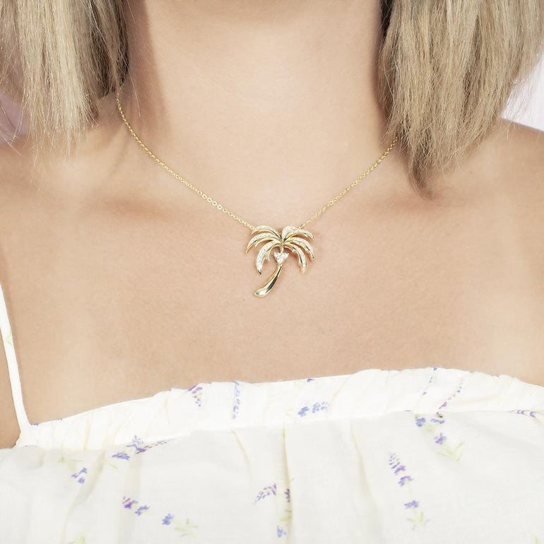 In this photo there is a model with blonde hair and a white shirt with flowers, wearing a yellow gold palm tree pendant with topaz gemstones.