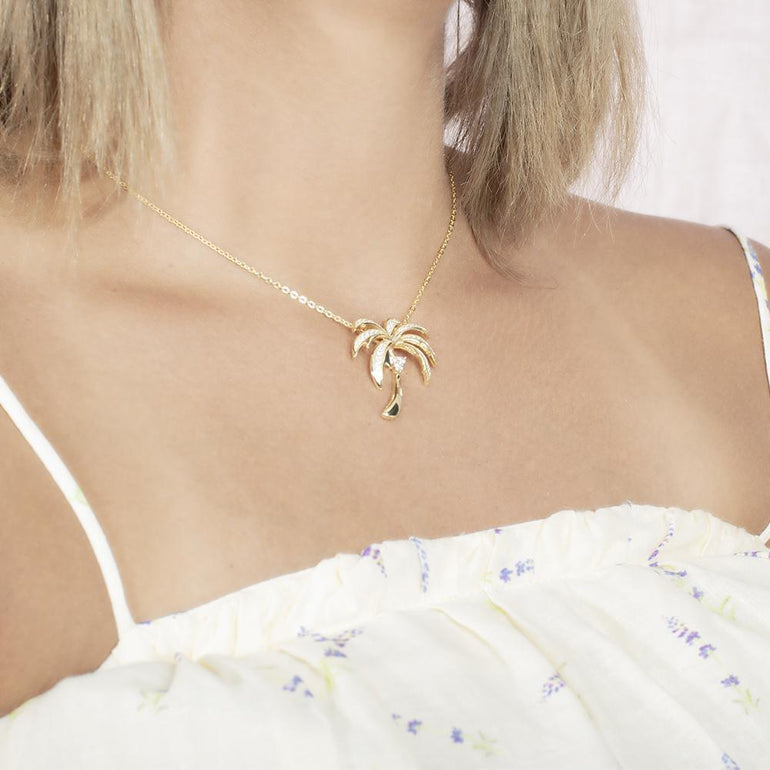 In this photo there is a model turned slightly to the right with blonde hair and a white shirt with flowers, wearing a yellow gold palm tree pendant with topaz gemstones.