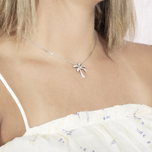 In this photo there is a model turned slightly to the right with blonde hair and a white shirt with flowers, wearing a white gold palm tree pendant with topaz gemstones.