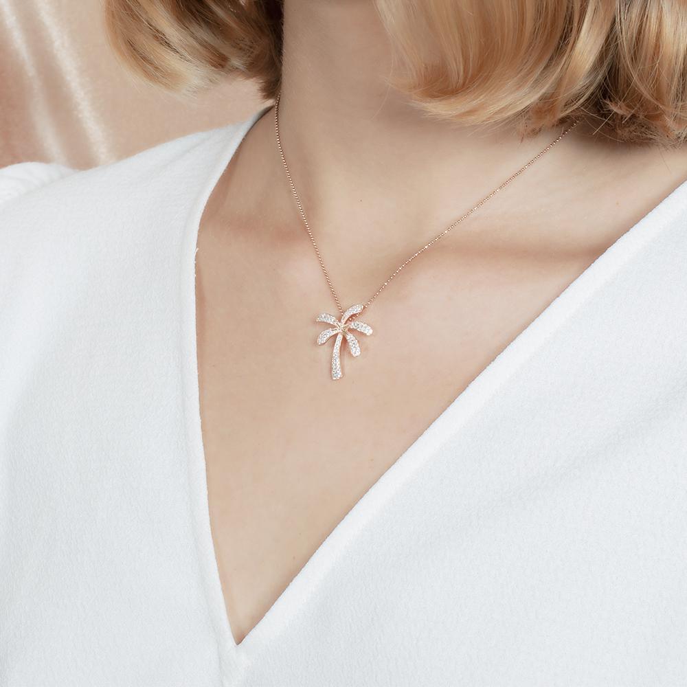 In this photo there is a model turned slightly to the left with blonde hair and a white shirt, wearing a rose gold palm tree pendant with topaz gemstones.