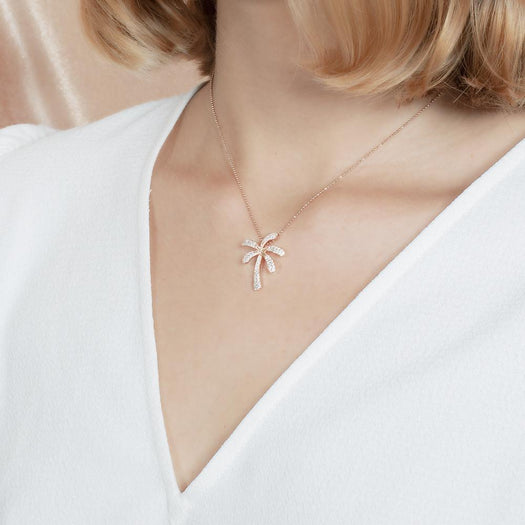 In this photo there is a model turned slightly to the left with blonde hair and a white shirt, wearing a rose gold palm tree pendant with topaz gemstones.