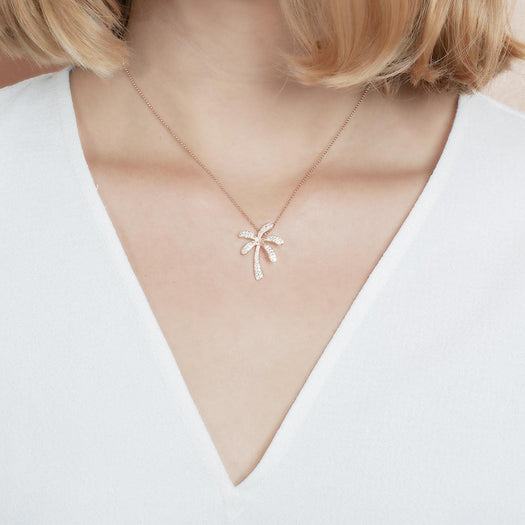 In this photo there is a model with blonde hair and a white shirt, wearing a rose gold palm tree pendant with topaz gemstones.