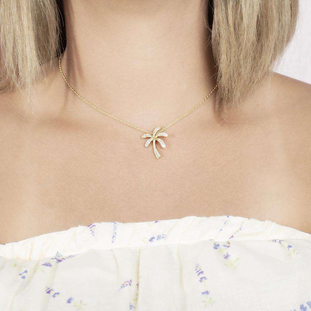 In this photo there is a model with blonde hair and a white shirt with flowers, wearing a yellow gold palm tree pendant with topaz gemstones.